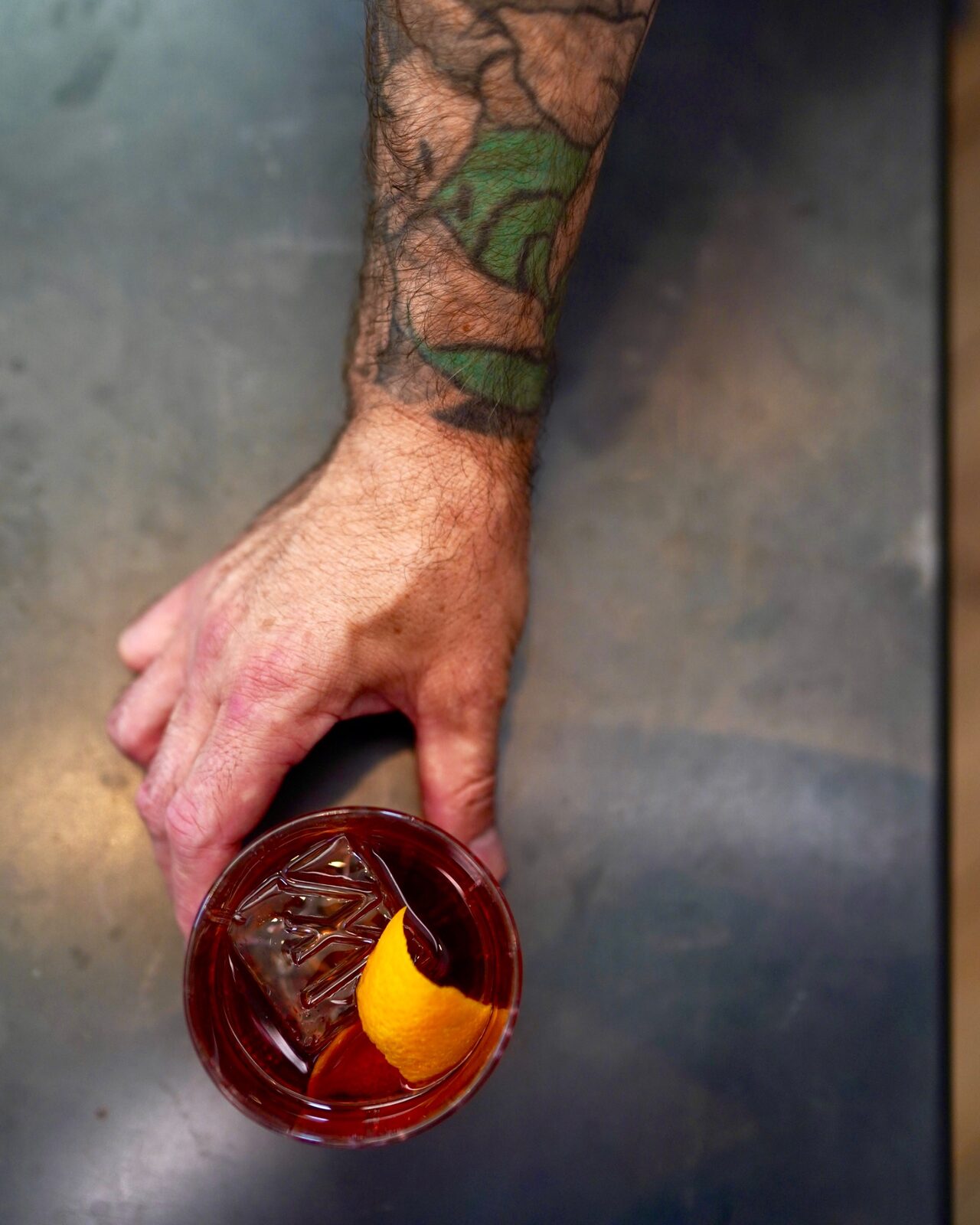 A tattooed hand holding an amber cocktail with an orange peel